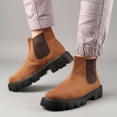 Brown Chelsea boots with black soles worn by a person on a wooden floor.