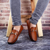 Brown loafers with gold buckles worn by a person against a brick wall.