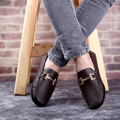 Brown loafers with gold buckles worn by a person sitting on a wooden stool against a brick wall.