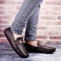 Brown loafers with gold buckles worn with gray jeans against a brick wall background