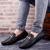 Black loafers with gold buckles worn with gray jeans against a brick wall.