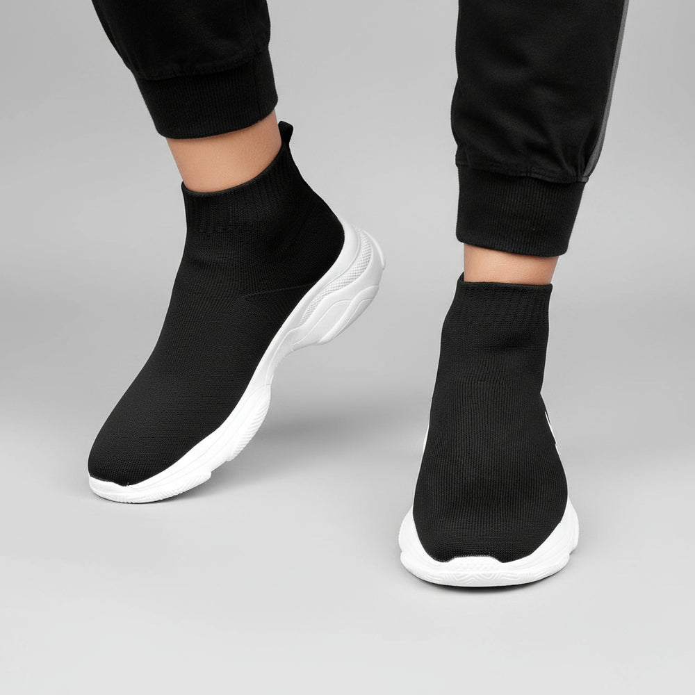 Black sock shoes with white soles worn by a person against a brick wall.