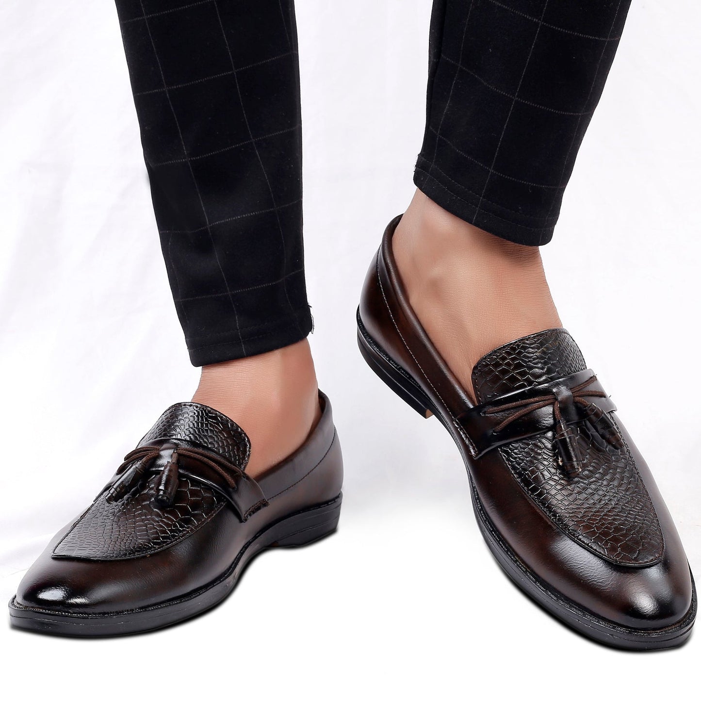 Brown leather loafers with tassels worn with black checkered pants on a white background