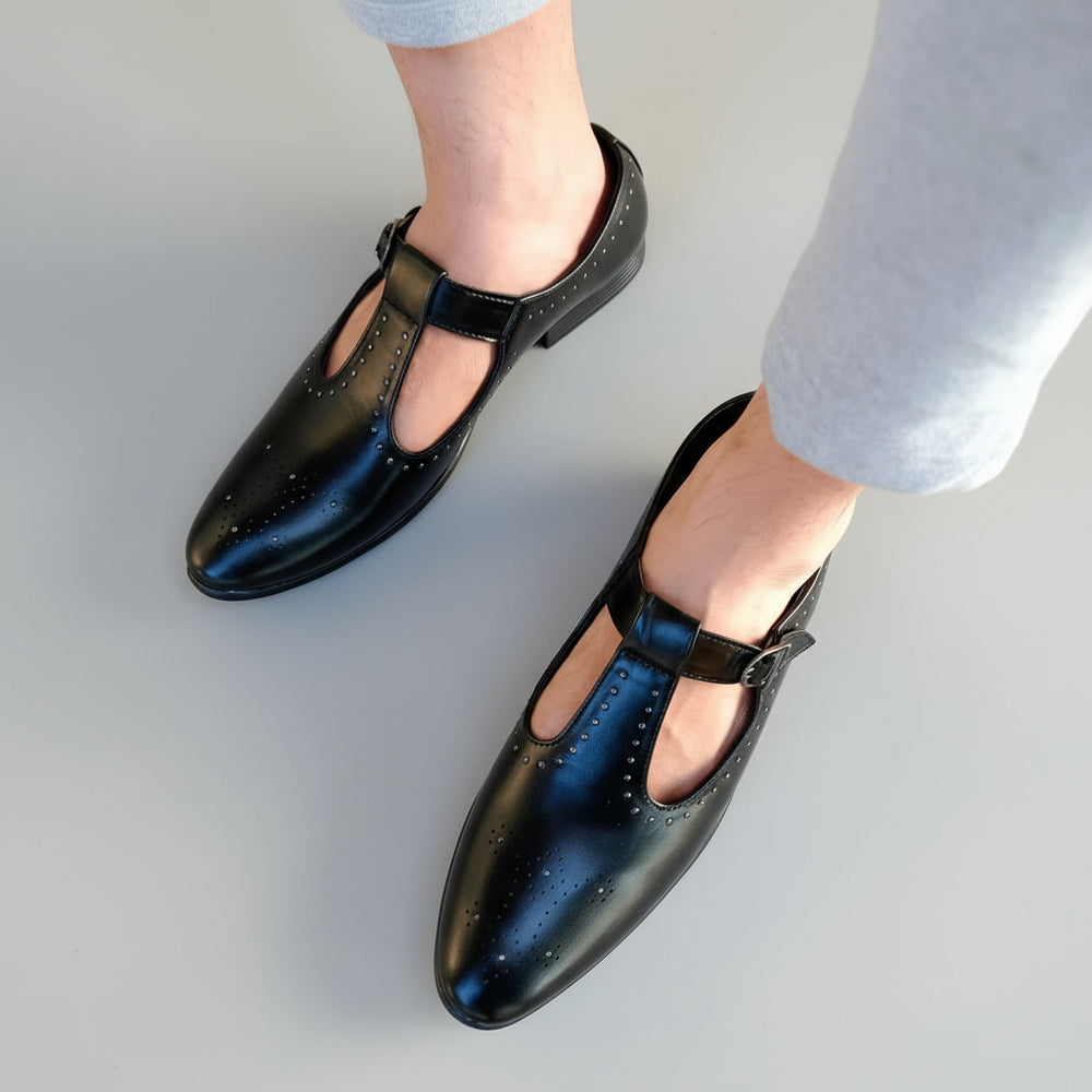 Black shoes with a strap worn by a person on a stone pavement.