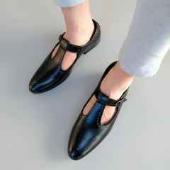 Black shoes with a strap worn by a person on a stone pavement.