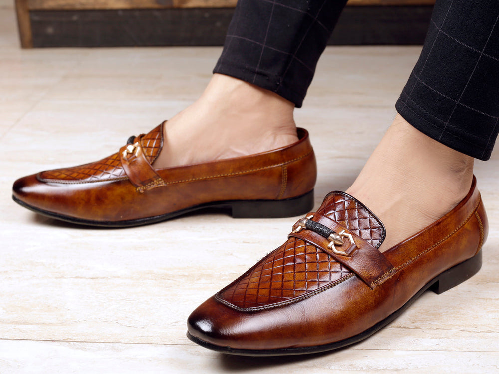 Brown leather loafers worn with dark pants on a light wooden floor.
