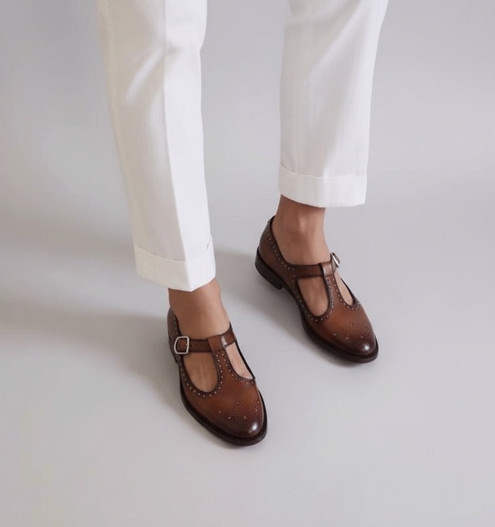 Brown shoes with a strap worn with white pants on a terrazzo floor.