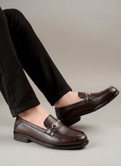 Brown loafers with gold accents worn by a person on a wooden floor.