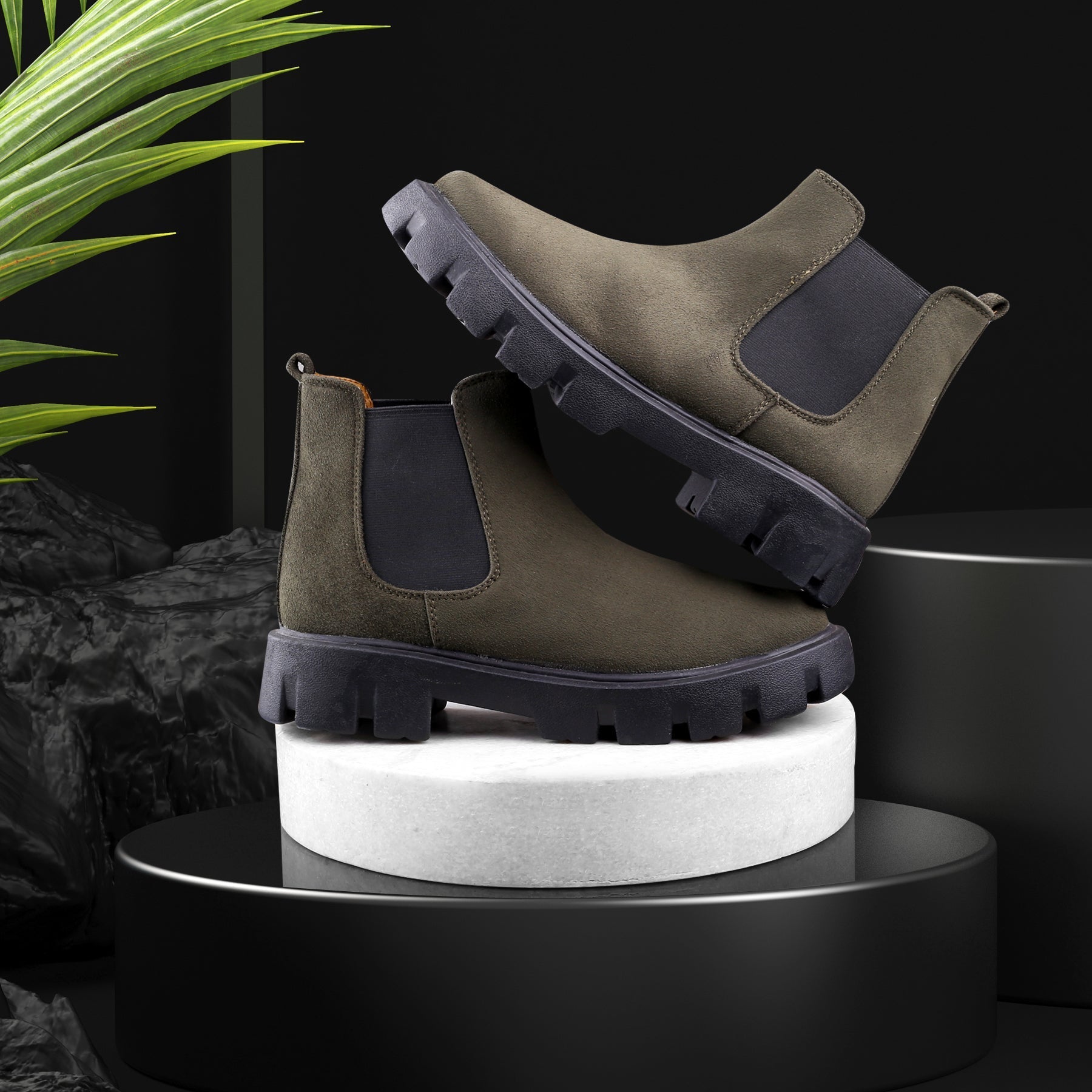 Pair of green suede boots with black soles on a white cylindrical platform against a dark background.