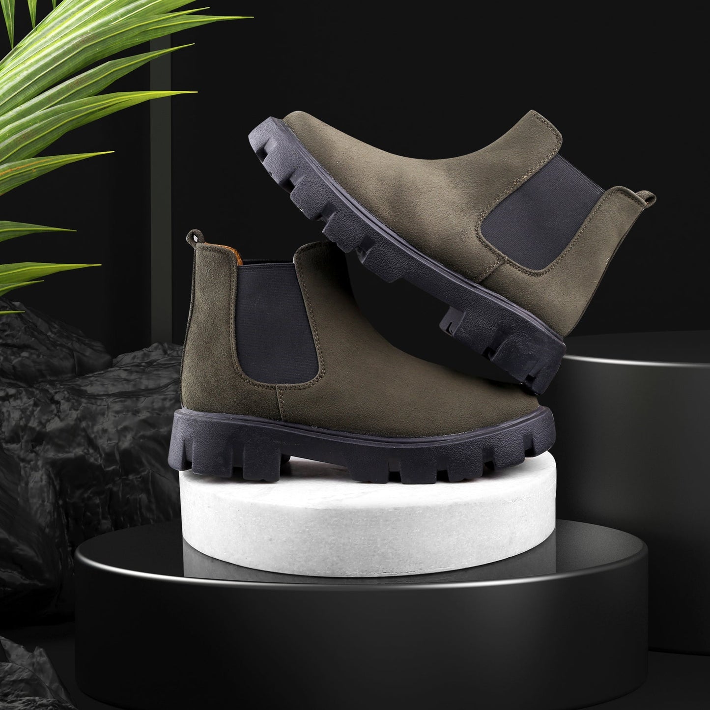 Pair of green suede boots with black soles on a white cylindrical platform against a dark background.