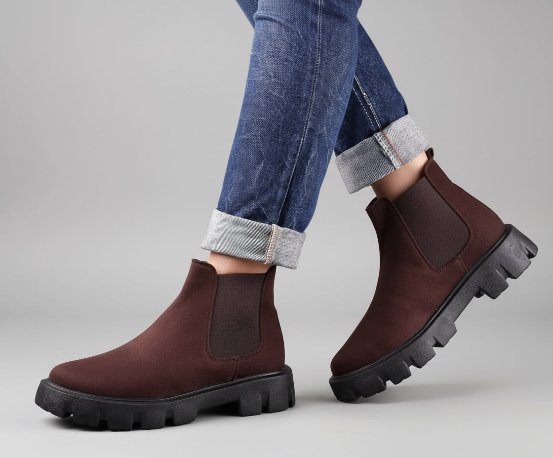 Brown Chelsea boots with black soles worn with rolled-up jeans on a wooden floor.