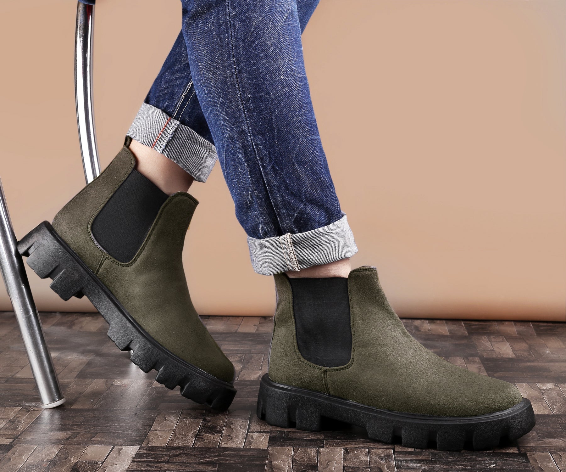 Green Chelsea boots worn with rolled-up jeans on a wooden floor.