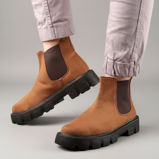 Brown Chelsea boots with black soles worn by a person on a wooden floor.