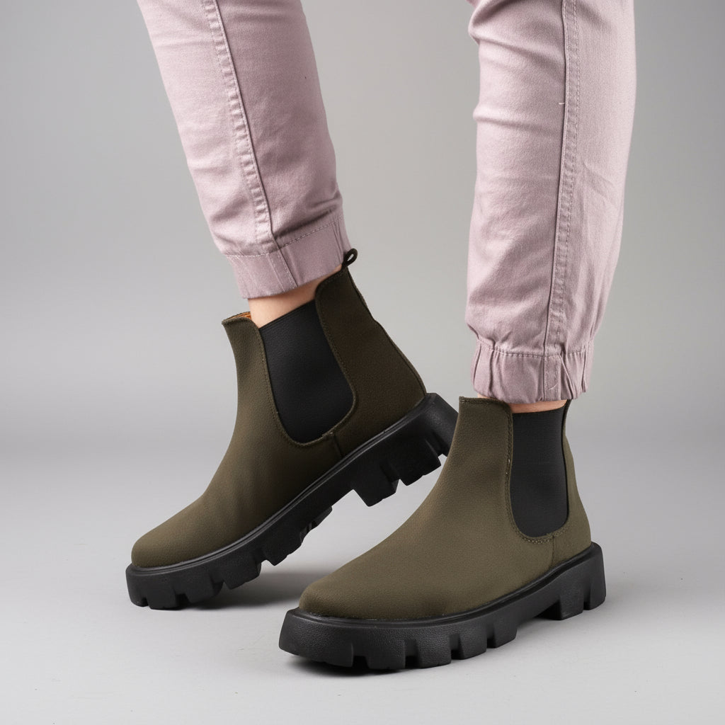 Green Chelsea boots with black soles worn by a person on a wooden floor.