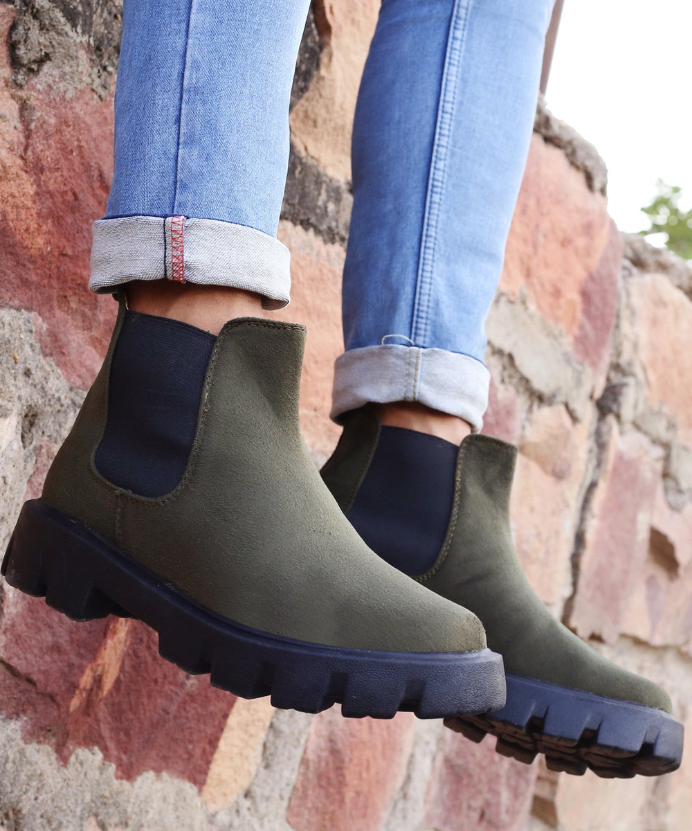 Green Chelsea boots with black soles worn with light blue jeans against a stone wall background.