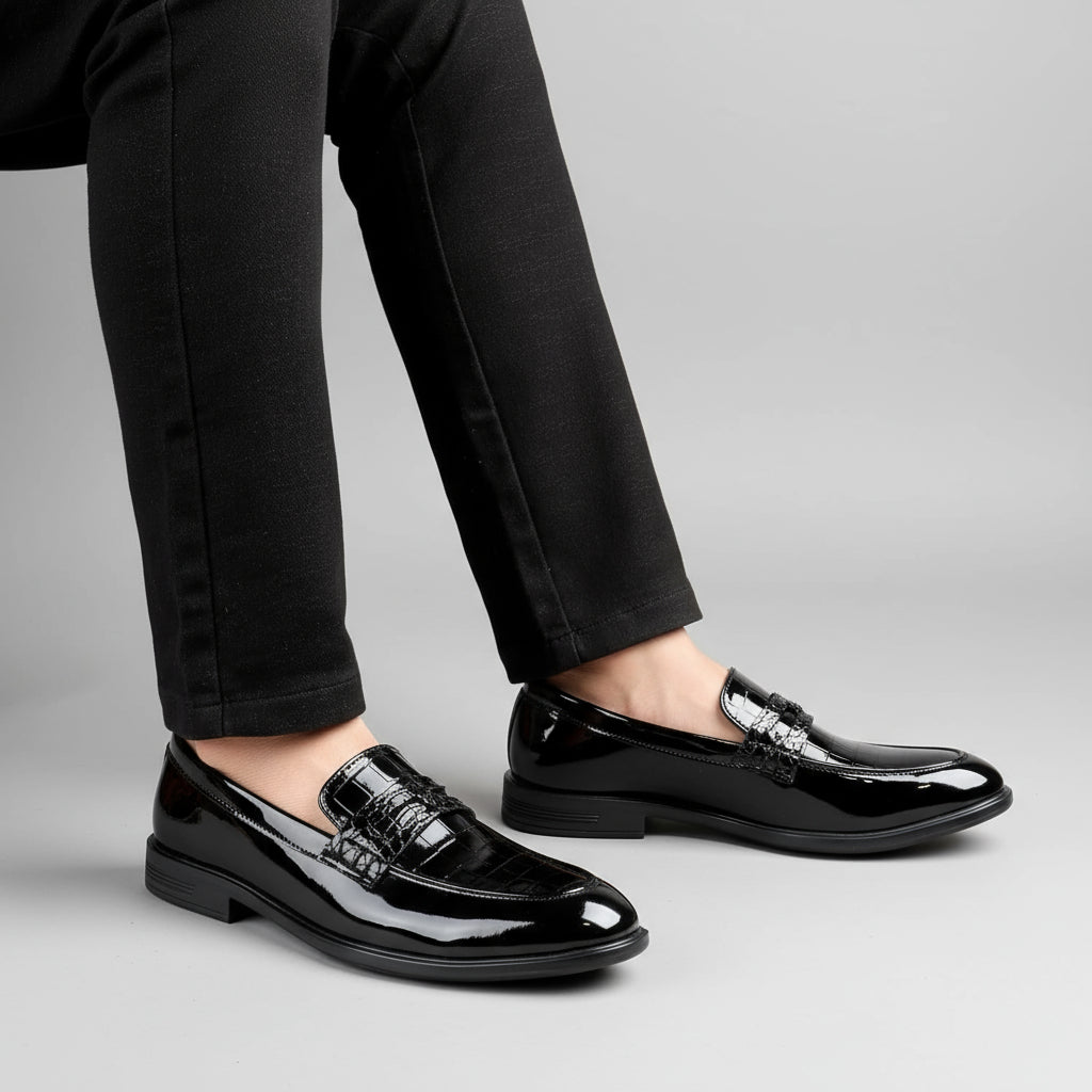 Black patent leather loafers worn with black pants on a wooden floor.