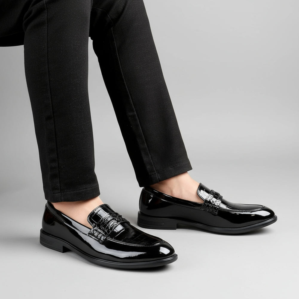 Black patent leather loafers worn with black pants on a wooden floor.