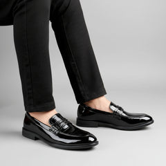Black patent leather loafers worn with black pants on a wooden floor.