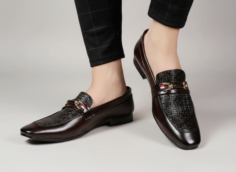 Brown leather loafers with decorative buckles worn by a person on a wooden floor.
