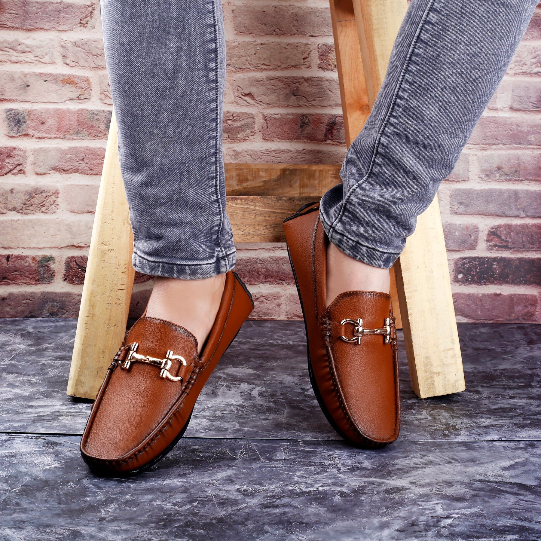 Brown loafers with gold buckles worn by a person against a brick wall.