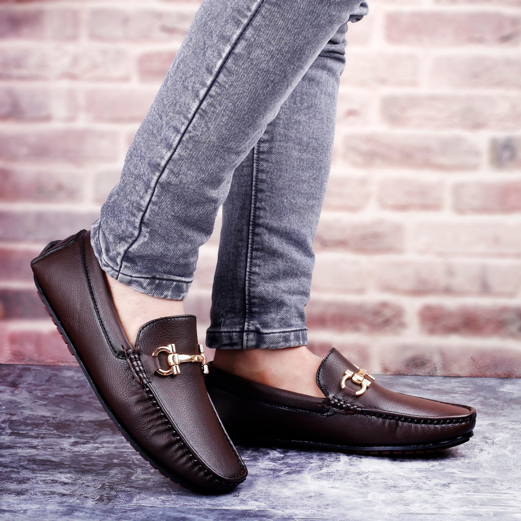 Brown loafers with gold buckles worn with gray jeans against a brick wall background