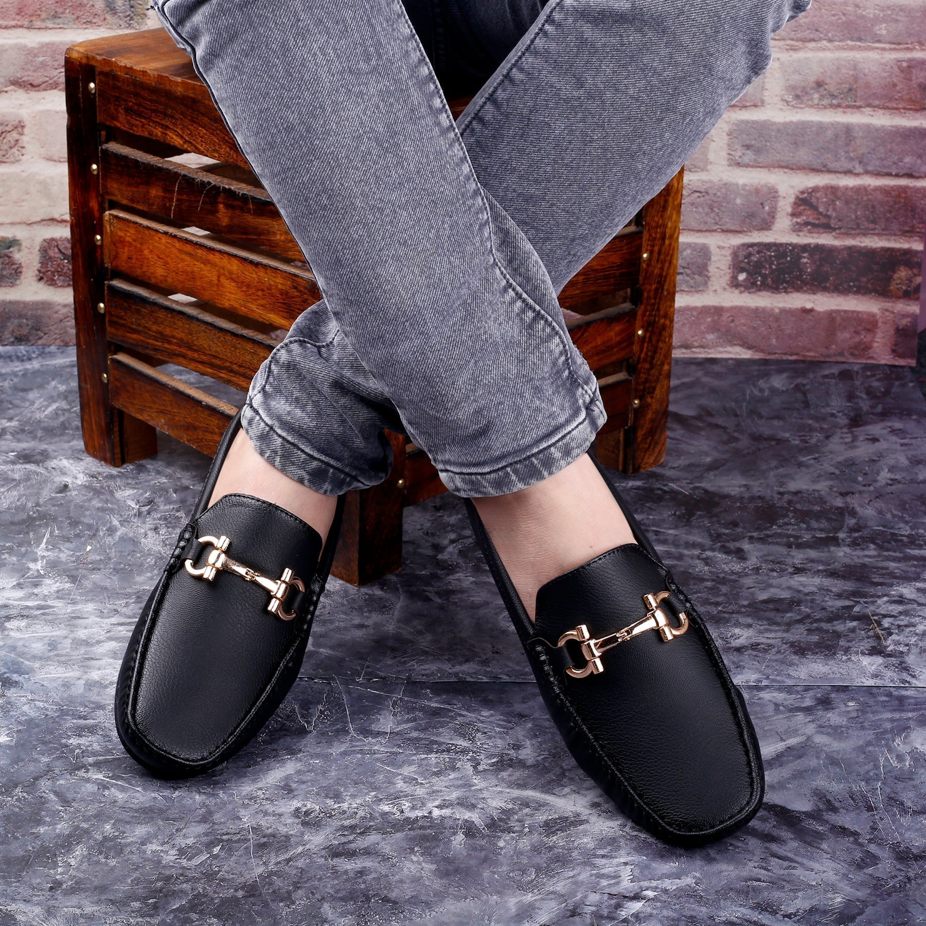 Black loafers with gold buckles worn by a person sitting on a wooden stool against a brick wall.