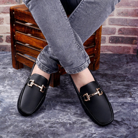 Black loafers with gold buckles worn by a person sitting on a wooden stool against a brick wall.