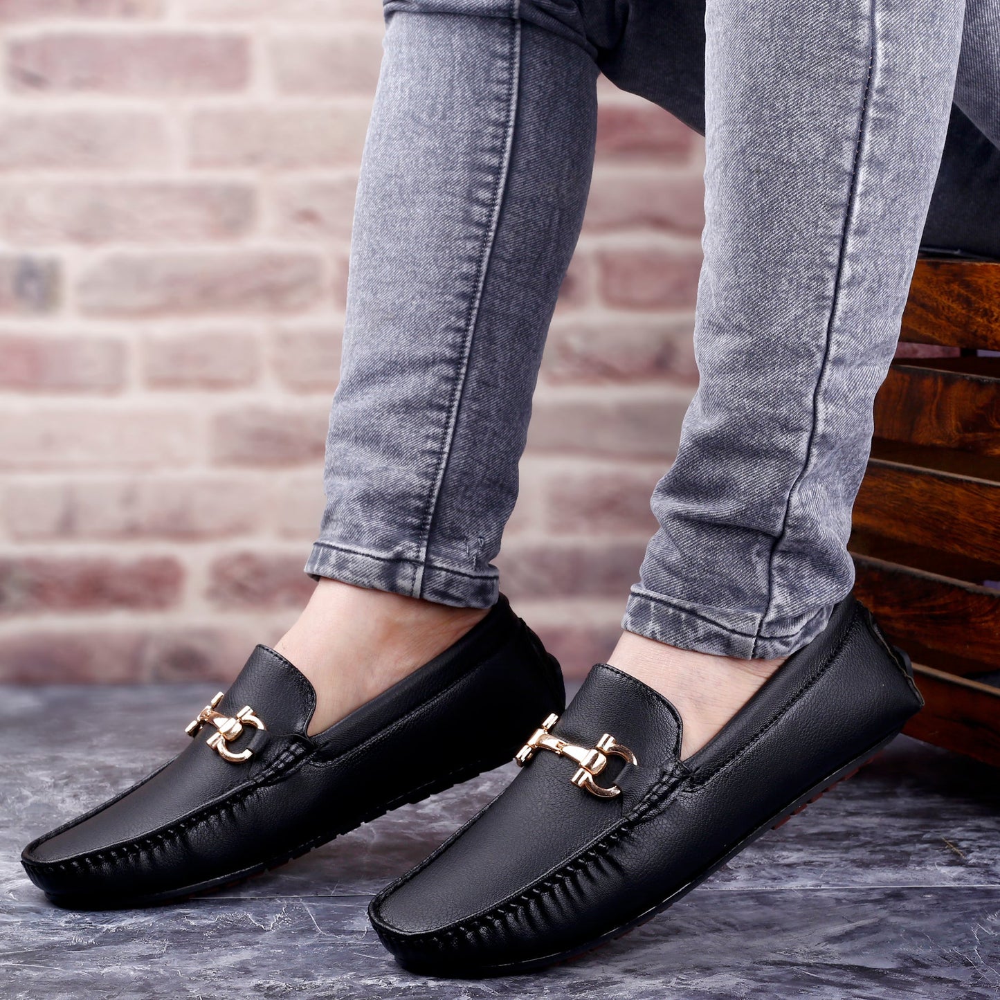 Black loafers with gold buckles worn with gray jeans against a brick wall.