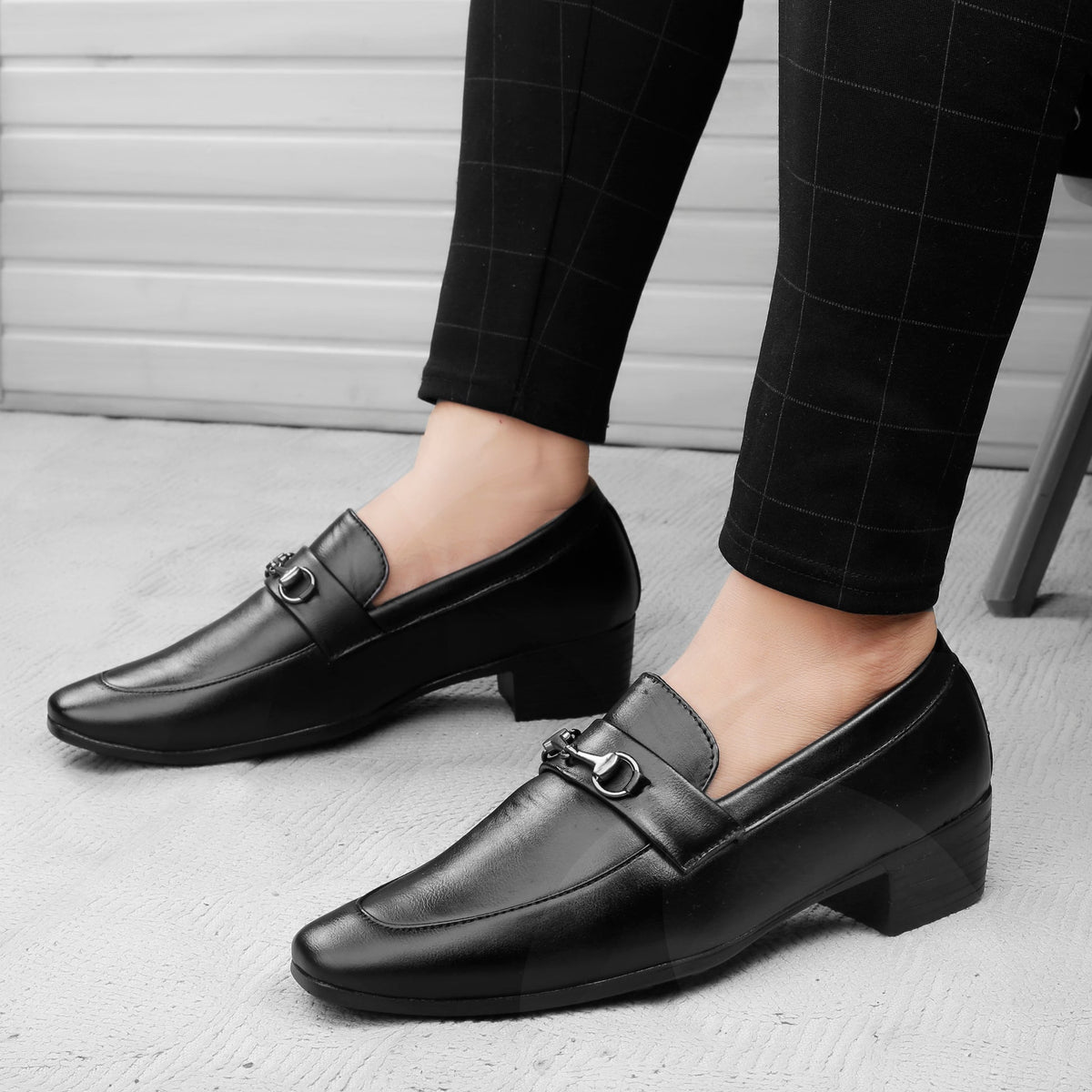 Black leather loafers worn with black checkered pants on a light gray floor.