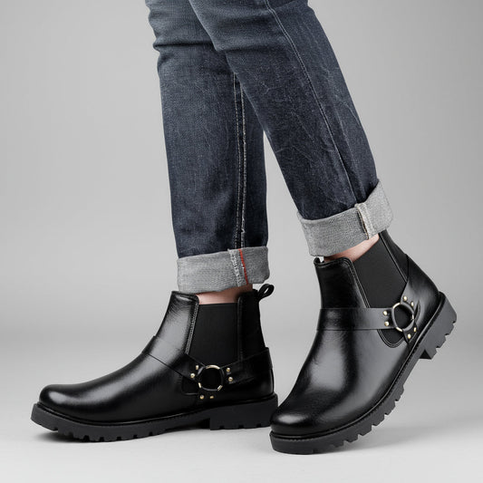 Black leather boots with gold buckles worn with rolled-up jeans against a wooden background