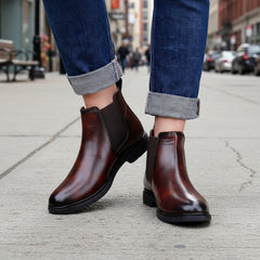 Brown leather Chelsea boots worn with rolled-up jeans on a wooden floor.