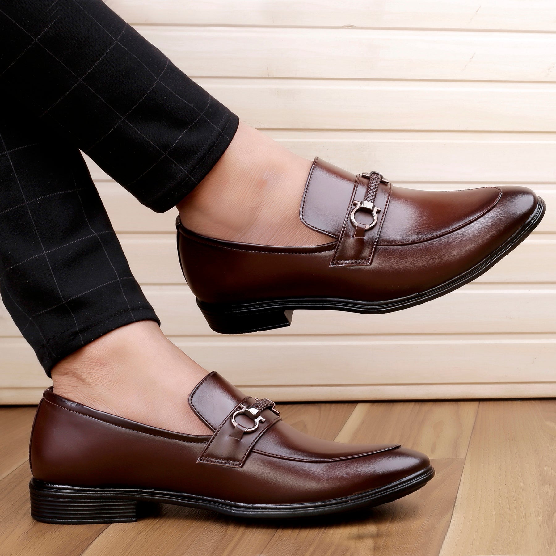 Brown leather loafers worn with black pants on a wooden floor.
