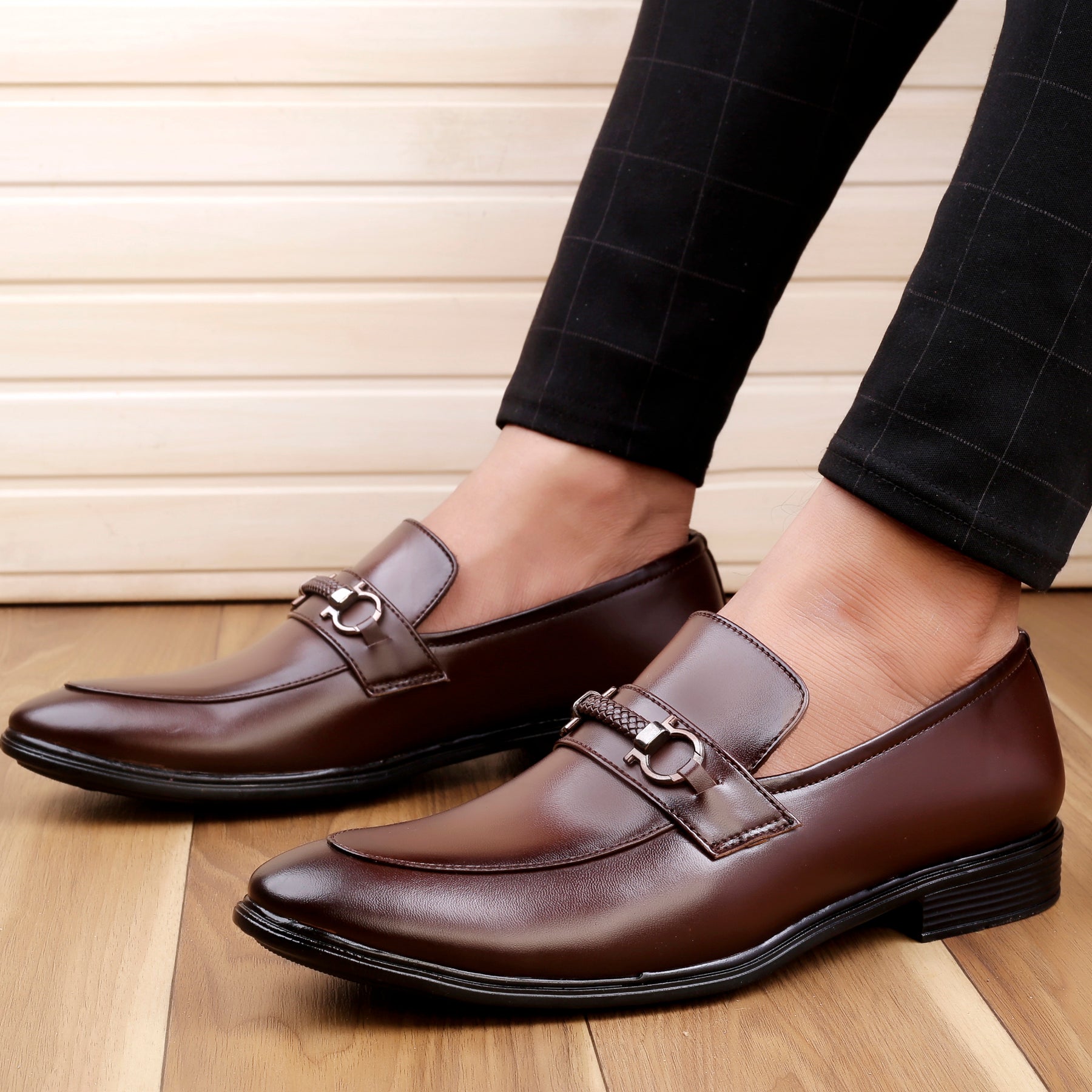 Brown leather loafers worn with black checkered pants on a wooden floor.