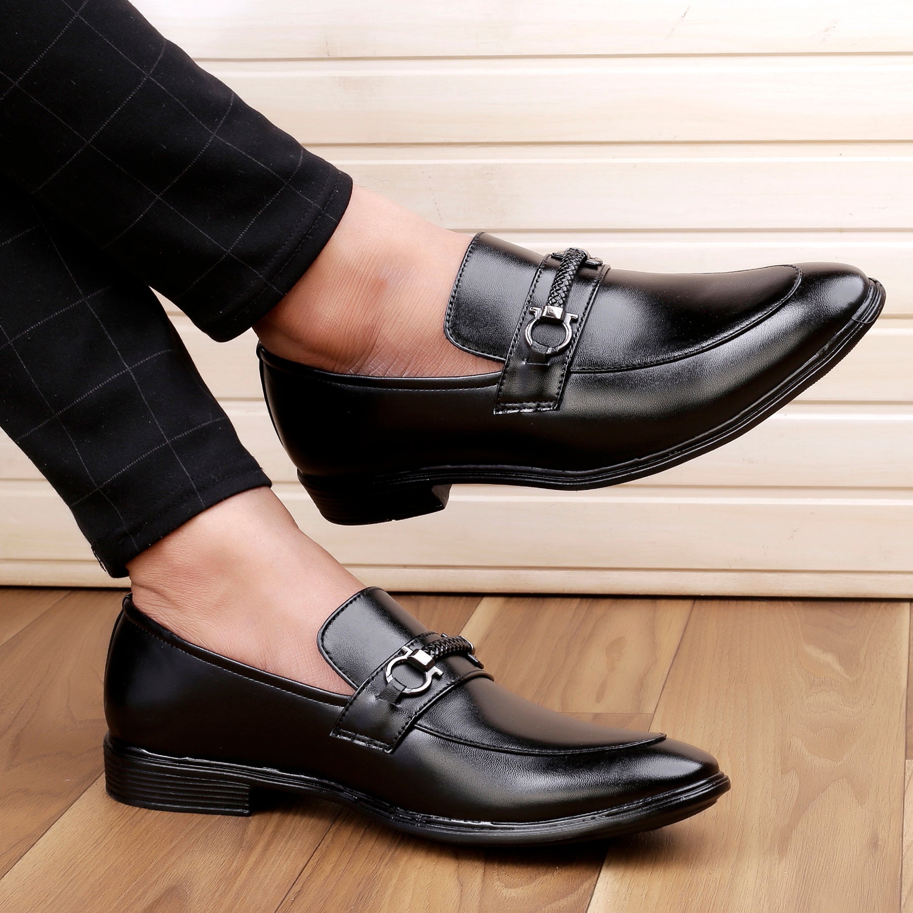 Black dress shoes worn with black pants on a wooden floor.