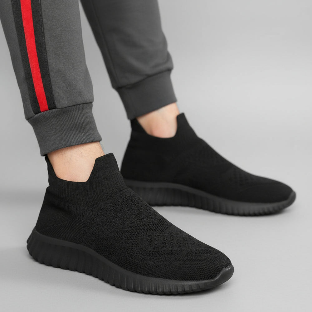 Black sock shoes worn with gray pants featuring a red stripe on a dark surface.