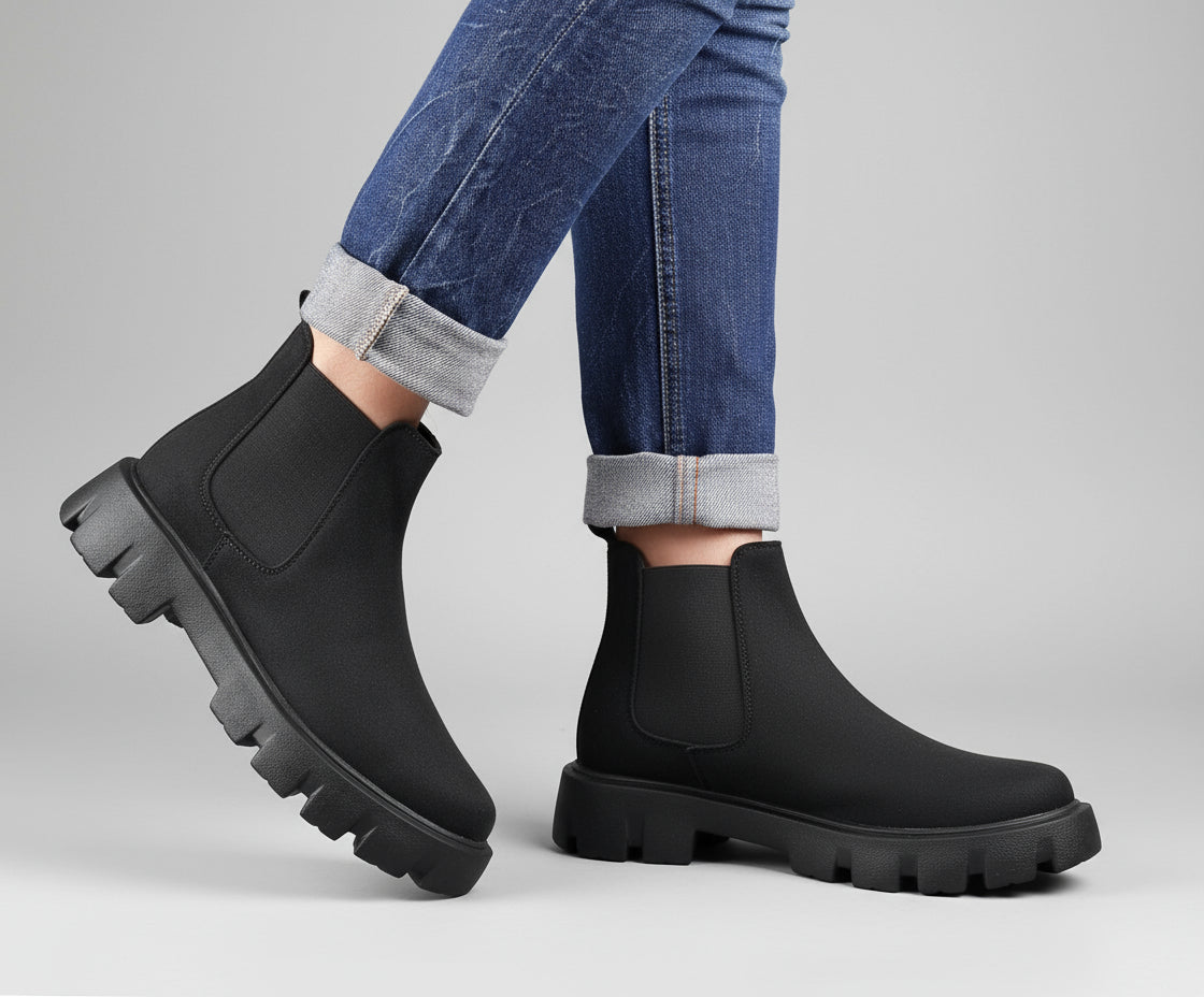 Black Chelsea boots worn with rolled-up jeans on a wooden floor.