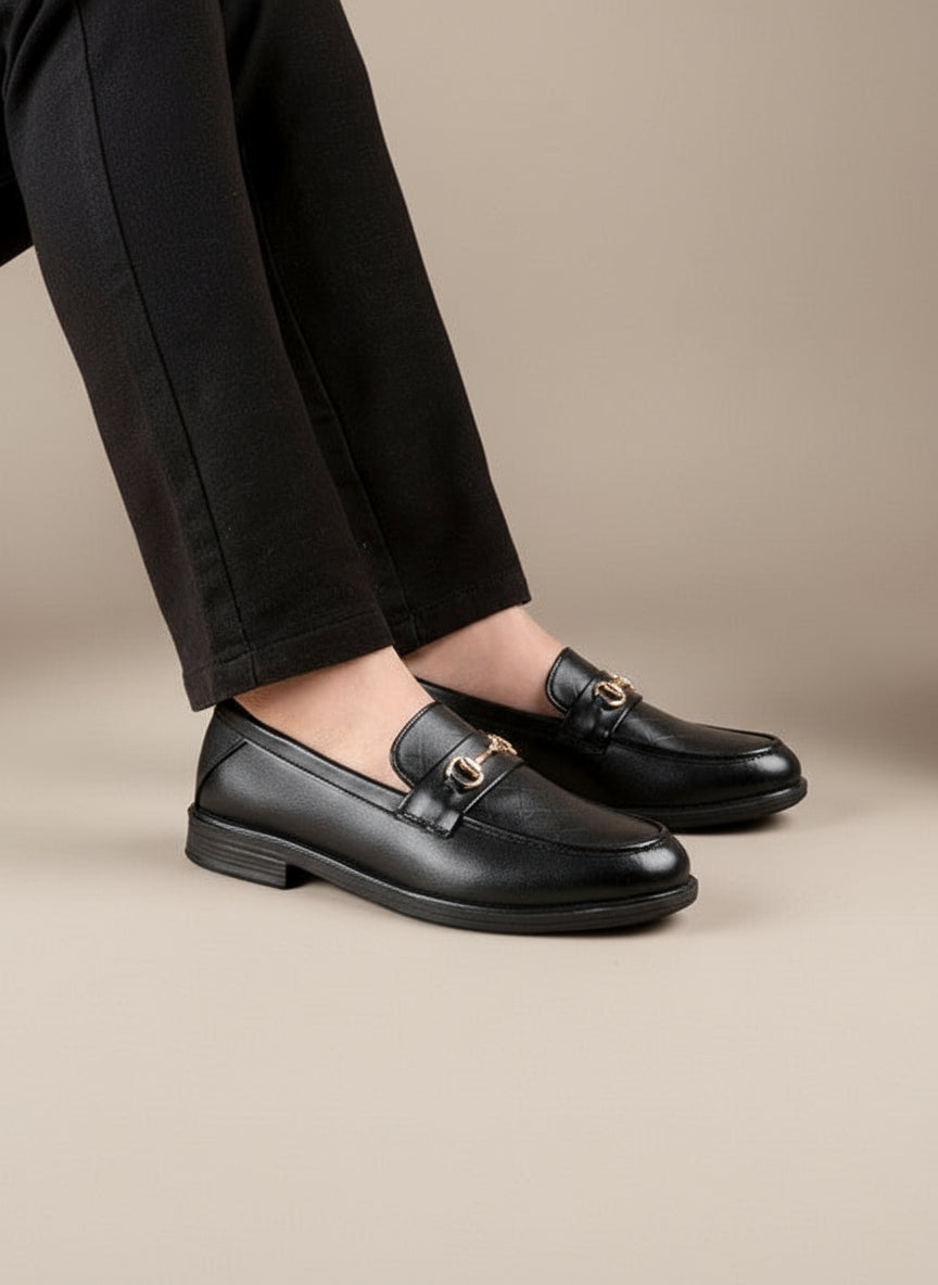 Black leather loafers with gold buckles worn by a person on a neutral background